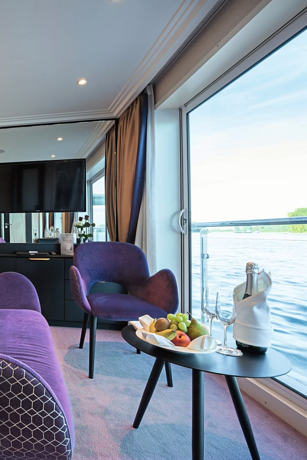 Doppelkabine auf der Excellence Countess mit lila Dekor, Samtstühlen, Obstkorb und Blick auf den Fluss.