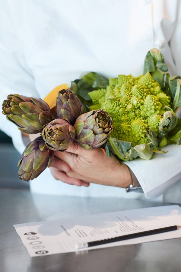 Koch hält Artischocken und Romanesco im Arm neben Rezeptblatt.