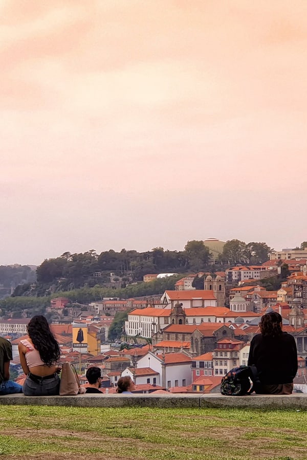 Menschen sitzen auf einer Wiese und blicken auf eine Stadtlandschaft bei Sonnenuntergang.
