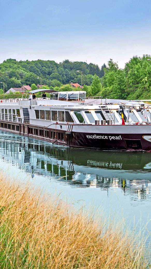dinnerkrimi-auf-dem-rhein-5.jpg Flusskreuzfahrtschiff auf einem ruhigen Kanal mit grünen Ufern.
