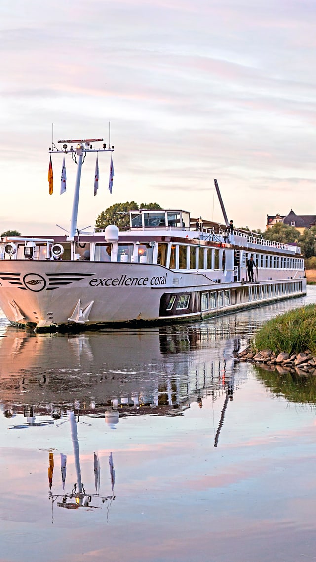 excellence-coral.jpg Flusskreuzfahrtschiff auf ruhigem Wasser nahe Ufer und Dorf.