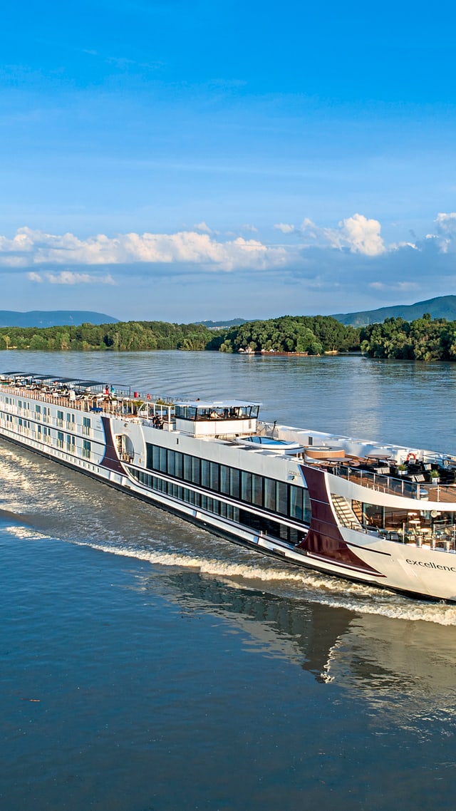 excellence-empress.jpg Flusskreuzfahrtschiff auf einem breiten Fluss mit bewaldetem Ufer.