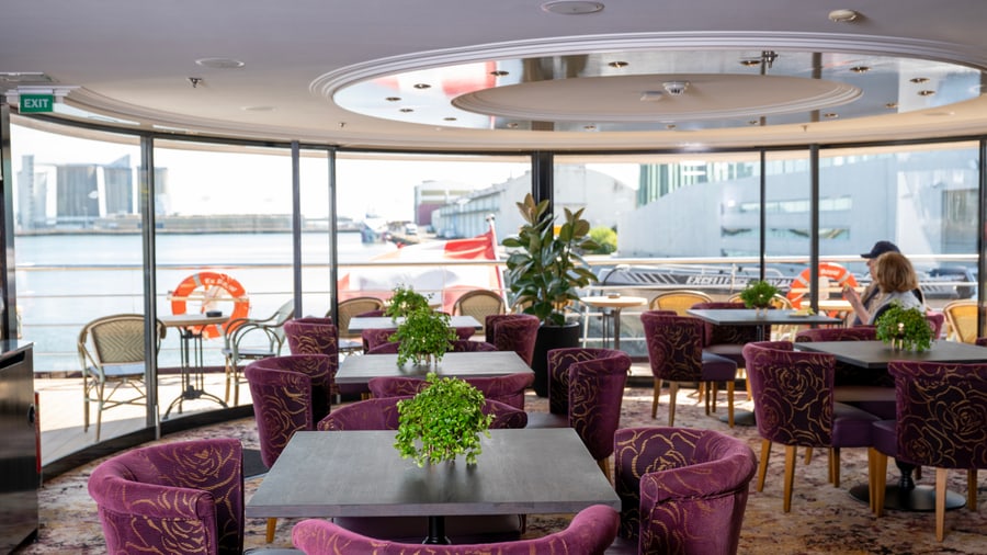 Restaurant mit lila Sesseln, Tischen und grossen Fenstern mit Blick auf das Wasser.