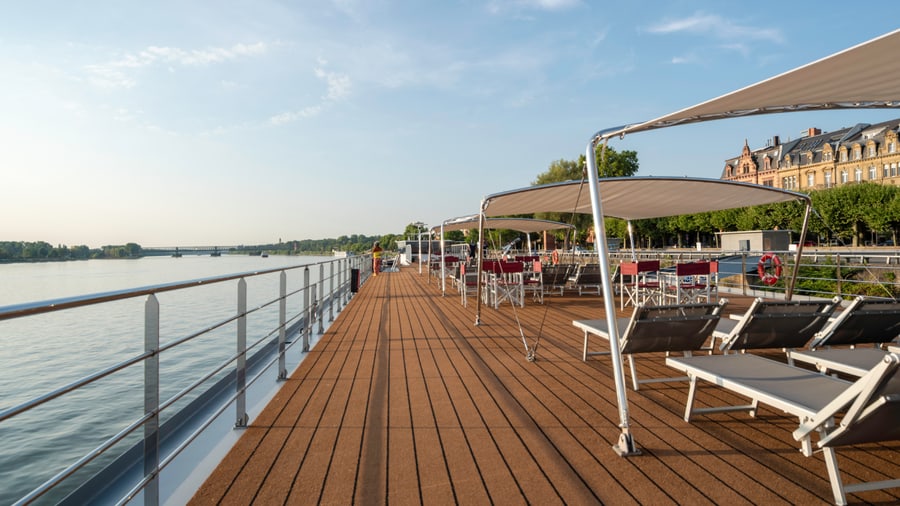 Deck eines Kreuzfahrtschiffs mit Liegestühlen und Sonnensegeln am Flussufer.