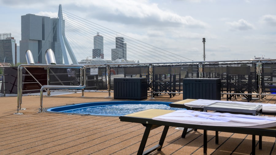 Deck eines Schiffes mit Pool und Liegestühlen, im Hintergrund eine Brücke und Wolkenkratzer.