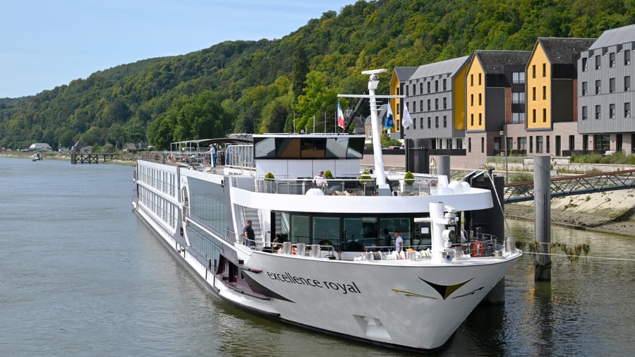 Flusskreuzfahrtschiff an einem Flussufer mit Hügeln und Gebäuden im Hintergrund.