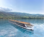 Flusskreuzfahrtschiff auf einem ruhigen Fluss vor bewaldeten Hügeln.