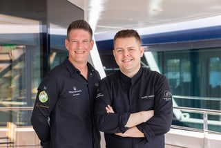 Zwei Männer in schwarzen Jacken stehen auf einem Schiffdeck.
