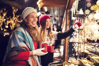 vier-staedte-maerchenhaft-magisch-10.jpg Zwei Frauen lächelnd auf einem Weihnachtsmarkt mit Tassen und Dekorationen.