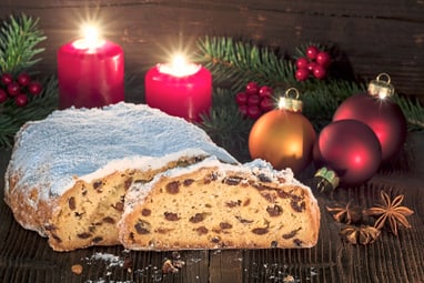 Ein angeschnittenes Stollenbrot mit roten Kerzen und Weihnachtsdekoration im Hintergrund.