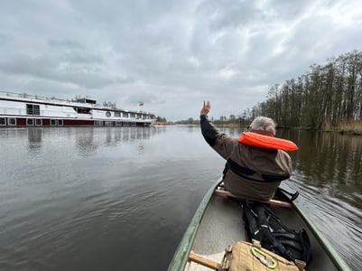 Person in einem Kanu winkt einem vorbeifahrenden Schiff zu, auf einem ruhigen Fluss.