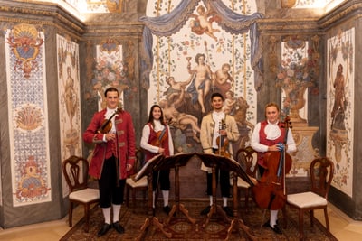 Vier Musiker in barocker Kleidung in kunstvoll dekoriertem Raum.