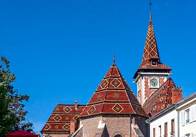 Gebäude mit gemusterten Dächern und Turm vor blauem Himmel.