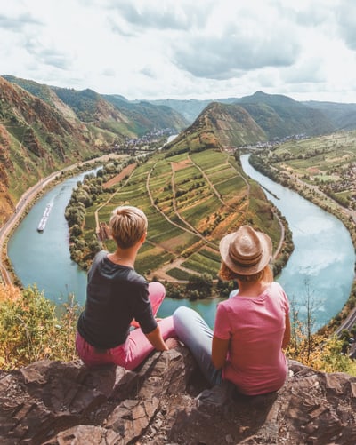 Zwei Personen sitzen auf Klippe mit Blick auf Fluss und Landschaft.