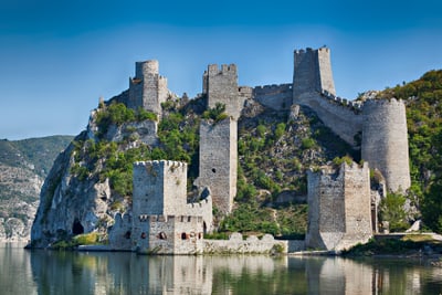 Mittelalterliche Burg am Flussufer mit bewachsenen Hügeln im Hintergrund.