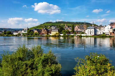 Flussufer mit Gebäuden im Hintergrund und bewaldetem Hügel.