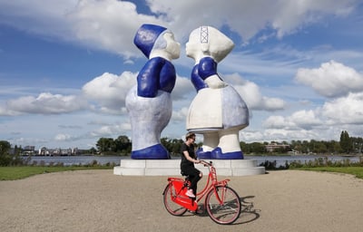 Person auf rotem Fahrrad vor grossen blauen Skulpturen.
