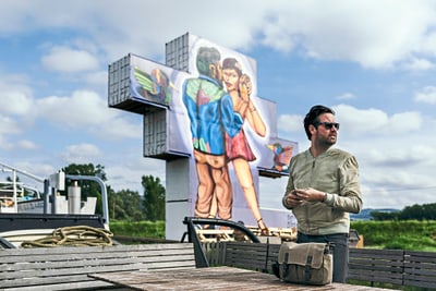 Mann mit Sonnenbrille vor grossem Wandgemälde im Freien, mit Wolkenhimmel.