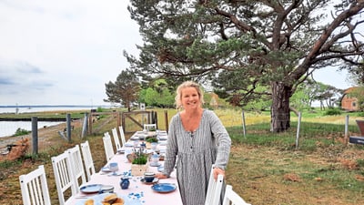 Frau steht neben langem gedeckten Esstisch im Freien am See.