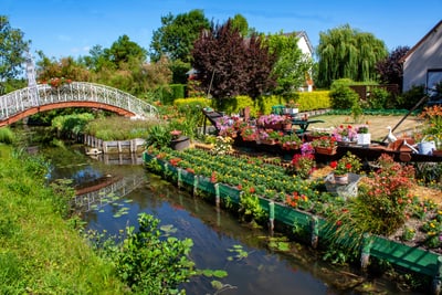 Garten mit Blumen am Fluss und einer Brücke im Hintergrund.