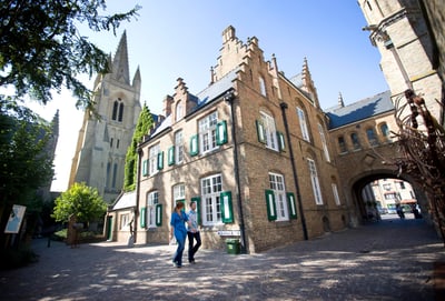 Mittelalterliche Gebäude mit Kirche im Hintergrund, zwei Personen gehen spazieren.
