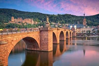 Historische Steinbrücke über einen Fluss vor einer malerischen Altstadt und Hügeln bei Sonnenuntergang.