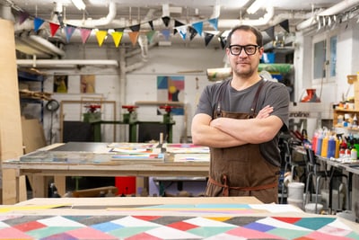 Mann mit Brille und Schürze steht in einer bunten Werkstatt.