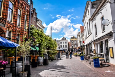 Strassenszene mit Menschen zwischen historischen Gebäuden und Pflanzen, blauer Himmel.