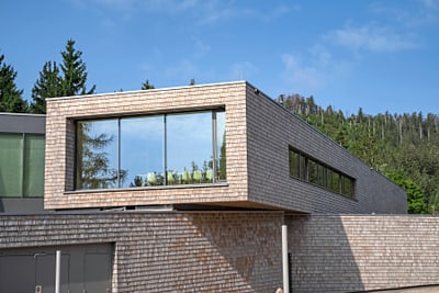 Modernes Gebäude mit holzverkleideter Fassade und grossen Fenstern vor einem bewaldeten Hintergrund.