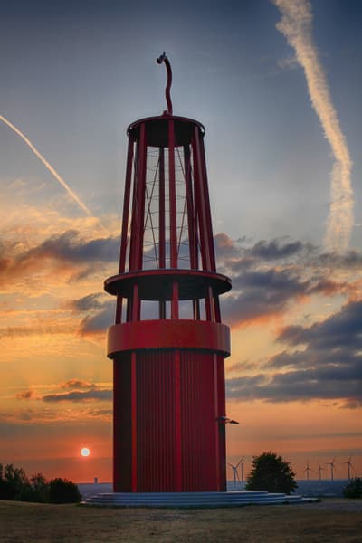 Grosse rote Skulptur vor einem Sonnenuntergangshimmel mit Wolken und Kondensstreifen.