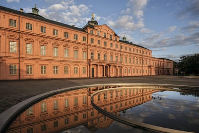 Historisches Gebäude mit rosa Fassade, reflektiert in einem Brunnen.