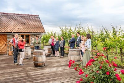 Gruppe von Menschen auf einer Terrasse mit Holzfässern und Rosen, vor Weinreben.
