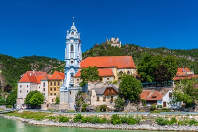 Historische Gebäude mit blauem Kirchturm am Fluss, umgeben von grünen Hügeln.