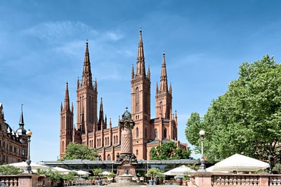 Historische Kirche mit hohen Türmen vor einem klaren Himmel, umgeben von Bäumen und Sonnenschirmen.