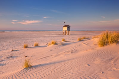 Sanddünen am Strand mit kleinem Pfahlhaus und blauem Himmel.