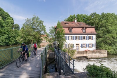 Zwei Personen auf Fahrrädern überqueren eine Brücke neben einem Fluss und einem Haus.