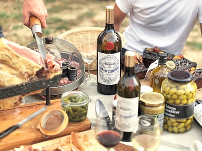 Person schneidet Fleisch auf einem Tisch mit Wein, Oliven und anderen Lebensmitteln.