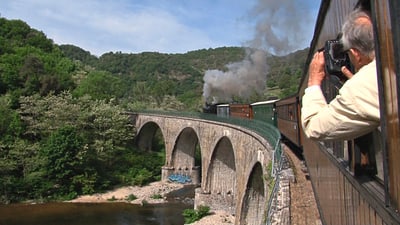 Zug fährt über eine steinerne Brücke, umgeben von Bäumen und Hügeln, mit einem fotografierenden Mann im Vordergrund.