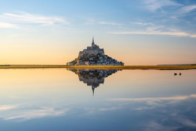 Burg auf Hügel, reflektiert im ruhigen Wasser, unter blauem Himmel.