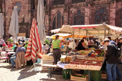 Marktszene mit Menschen, Verkaufständen und Schirmen vor einem alten Gebäude.