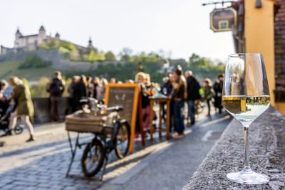 Glas Wein auf einer Mauer, Menschen gesellig an einem Tisch im Freien, historische Gebäude im Hintergrund.