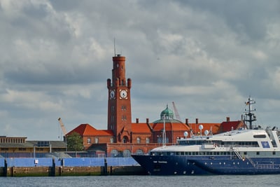 Grosses Gebäude mit Uhrturm und rotem Dach, im Vordergrund ein grosses Schiff.