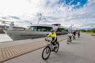 Personen fahren auf Fahrrädern entlang eines Flusses mit einem grossen Schiff im Hintergrund.