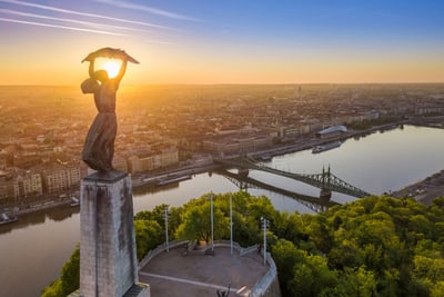 Freiheitsstatue auf Hügel mit Fluss und Stadt im Sonnenuntergang.