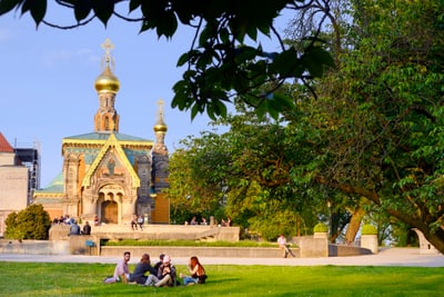 Menschen sitzen auf Rasen vor russisch-orthodoxer Kapelle in sonnigem Park.