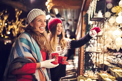 vier-staedte-maerchenhaft-magisch-10.jpg Zwei Frauen lächelnd auf einem Weihnachtsmarkt mit Tassen und Dekorationen.