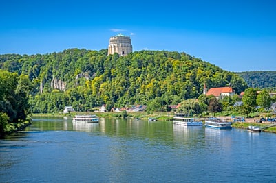 Ruhiger Fluss mit Schiffen, Hügel und Gebäude im Hintergrund.