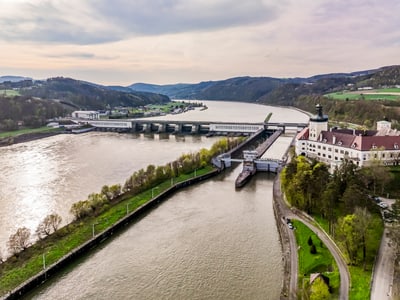 Luftaufnahme eines Flusses mit Dämmen und Gebäuden am Ufer, umgeben von grüner Landschaft.