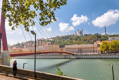 Flussufer mit Brücke, Hügel mit Gebäuden und Turm im Hintergrund.