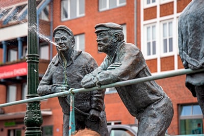 Bronzefiguren zweier Männer, einer spuckt Wasser.